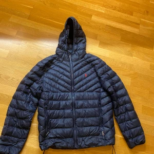 Navy dunjacka från Polo Ralph Lauren - Snygg svart dunjacka från Polo Ralph Lauren med huva och dragkedja framtill. Jackan har quiltad design, två fickor med dragkedja och röd logga på bröstet. 