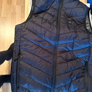 Svart värmeväst med USB och dragkedja - Svart dunväst med värmefunktion som aktiveras med knapp på bröstet. Västen har dragkedja framtill, två fickor med dragkedja och ett glansigt, reflekterande innerfoder. USB-kabel för laddning ingår. Perfekt för kalla dagar när du vill hålla dig varm och bekväm.