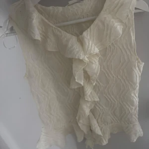Krämvit blus med volang från Gina Tricot - Supersöt krämvit blus från Gina Tricot med volangdetaljer längs kragen och framtill. Blusen är ärmlös och har ett vågigt, strukturerat tyg!💗