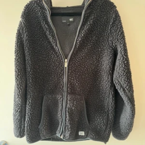 Mörkgrå teddyjacka med huva  - Mysig mörkgrå teddyjacka från lager 157 med huva och dragkedja framtill. Jackan har två stora fickor och är tillverkad i fluffigt teddy-material som håller dig varm. Perfekt för kyliga dagar och har en avslappnad passform.