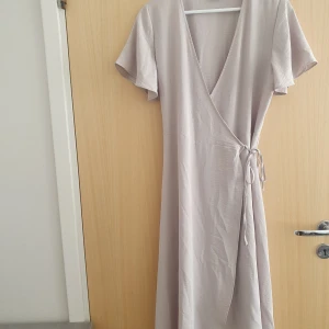 Ljus omlottklänning med knytband - En stilren ljus långklänning med omlottmodell och knytband i midjan. Klänningen har korta ärmar och v-ringad hals, vilket ger en luftig och avslappnad känsla. Klänningen är i storlek 40 men passar även de som har storlek 36. Klänningen köptes för 400 kr men då den är använd ett fåtal gånger kan jag tänka mig gå ner i pris