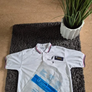 Vit pikétröja från Ralph Lauren M - Vit pikétröja från Ralph Lauren med klassisk krage och korta ärmar. Tröjan har tunna röda och blå ränder på krage och ärmslut samt broderad logga på bröstet. Tillverkad i bomull för en skön och luftig känsla. Perfekt för en clean och sportig stil.