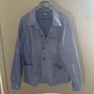Blå overshirt från Ralph Lauren - Snygg blå overshirt från Polo Ralph Lauren med tre stora fickor framtill och knäppning med svarta knappar. Jackan har en avslappnad passform och är tillverkad i bomull. Perfekt för lager-på-lager och har coola vita färgstänk för en unik look. Skick 9/10 