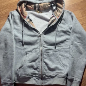 Grå hoodie från Burberry med dragkedja - Säljer en grå hoodie från Burberry med klassiskt rutigt foder i huvan och broderad logga på bröstet. Tröjan har dragkedja, snörning i huvan och stora fickor framtill. Mjuk insida och riktigt snygga detaljer. Perfekt för dig som gillar streetstyle.