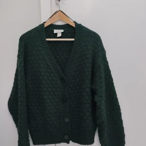 Mörkgrön stickad kofta från Monki - Mysig mörkgrön stickad kofta från Monki i storlek S. Koftan har ett bubbligt mönster, djup v-ringning och stora knappar framtill. Långa ärmar och ribbade muddar ger en chill och loose vibe. Perfekt att slänga på sig när det är kallt ute.