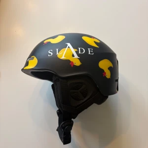 Skidhjälm med gula ankor från SLADE - Cool svart hjälm från SLADE med unikt tryck av gula ankor och vit logga. Hjälmen har justerbart hakband och ventiler på sidorna. Du får buffen på köpet!  Någon ynka repa på vänstra sidan men annars i gott skick! Jag säljer även en matchande skidjacka till om någon är intresserad 🥰