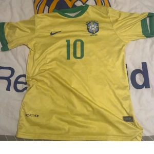 Gul Brasilien fotbollströja Nike - Säljer en klassisk gul Brasilien fotbollströja från Nike med gröna detaljer och nummer 10 på bröstet. Tröjan har korta ärmar, grönt krage och är tillverkad i lätt och ventilerande Dri-FIT material. Perfekt för dig som älskar fotboll och vill sticka ut på planen.
