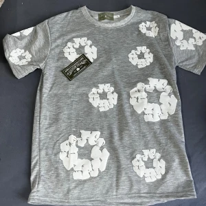 Grå t-shirt med vita blommor Denim Tears - Gråmelerad t-shirt från Denim Tears i bomull med unika vita blommönster tryckta över hela plagget. Klassisk passform och rund halsringning. Perfekt för dig som vill sticka ut med en cool och modern stil.