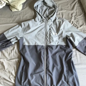 Grå vindjacka från Under Armour - Snygg tvåtonad grå vindjacka från Under Armour med huva och hel dragkedja. Jackan har en bröstficka med dragkedja, justerbar huva och reflekterande detaljer på axlarna. Perfekt för träning utomhus och har en sportig vibe.