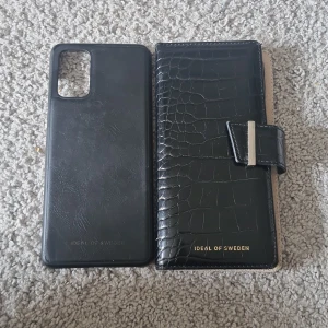 Svart plånboksfodral Ideal of Sweden - Snyggt svart plånboksfodral från Ideal of Sweden med krokopräglad yta och magnetlås. Fodralet har plats för kort och mobilen, och är gjort i skinnimitation. Perfekt för dig som vill ha både stil och funktion i ett.