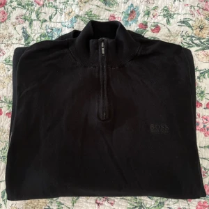  Hugo Boss half zip tröja - Snygg svart tröja från Hugo Boss med half zip och hög krage. Tillverkad av fin italiensk ull, perfekt för dig som gillar stilrena och klassiska plagg. Diskret Boss-logga på bröstet och ribbade detaljer vid halsen. Storlek S. 