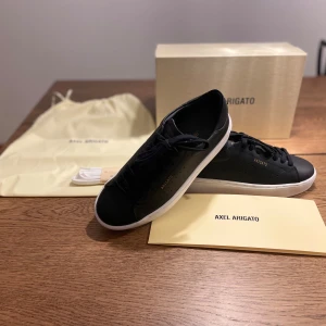 Svarta sneakers från Axel Arigato - Stilrena svarta sneakers från Axel Arigato i skinn med vit sula och diskret guldfärgad logga på sidan. Klassisk låg modell med svarta snören, perforerade detaljer och rund tå. Levereras med originalkartong, dustbag och extra vita skosnören.