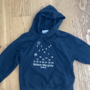 Maison Margiela svart hoodie med tryck - Svart hoodie från Maison Margiela med ikoniskt siffror-tryck och logga på bröstet. Klassisk huva och långa ärmar, tillverkad i mjuk bomull. Snygga vita stygn på ryggen som ger den där Margiela-viben. Perfekt för dig som gillar streetwear med edge.