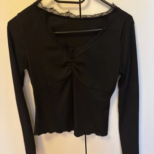 Svart långärmad topp med spets - Svart långärmad topp med v-ringning och spetsdetaljer längs halsringningen. Toppen har rynk framtill och en croppad passform med vågig nederkant. Perfekt för dig som gillar stilrena och trendiga plagg.