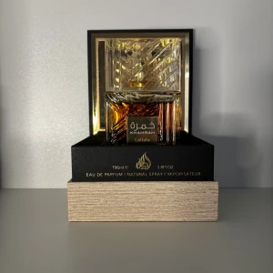 Khamrah Eau de Parfum sampel - Khamrah från Lattafa är en lyxig parfym i en elegant, fyrkantig glasflaska med guldiga detaljer och bärnstensfärgad vätska. Flaskan har ett mönstrat lock och levereras i en exklusiv svart och guldig förpackning. Volymen är 100 ml.