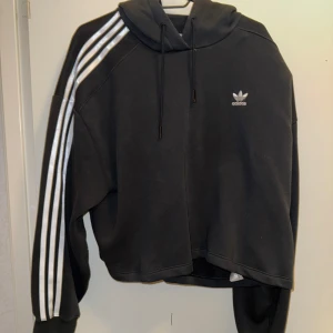 Svart Adidas hoodie med vita ränder - Svart hoodie från Adidas med klassiska vita tre ränder längs ärmarna och liten logga på bröstet. Tröjan har huva med snörning och är i en loose passform. Perfekt för en sportig och chill stil.