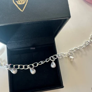 Pärlhalsband Silver - Snyggt halsband i silverfärgad metall med grova länkar och hängande droppformade pärlor. Perfekt för att ge din outfit en cool och trendig touch. Kommer i svart ask med gulddetalj på locket. Passar dig som gillar statement-smycken och pärlor.