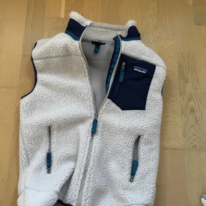 Beige fleeceväst från Patagonia - Säljer en beige fleeceväst från Patagonia i storlek S. Västen har en mjuk och fluffig yta, marinblå detaljer och tre fickor med dragkedja. Perfekt med hög krage och coolt bröstficka med logga. Snygg att slänga över en hoodie eller t-shirt.
