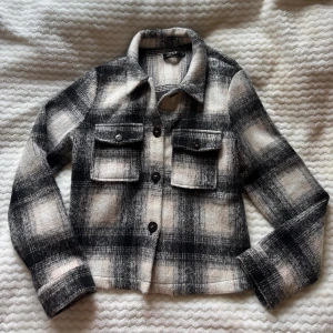 Rutig kappa från ONLY  - Snygg rutig kappa från ONLY i svart, beige och grå toner. Jackan har klassisk krage, två bröstfickor med knappar och stängs med stora svarta knappar framtill. Perfekt för lager-på-lager och ger en cool vibe till din outfit.