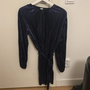 Mörkblå plisserad klänning NLY Trend - Snygg mörkblå plisserad kortklänning från NLY Trend. Klänningen har långa ärmar, rund hals och ett matchande knytband i midjan. Materialet är glansigt och ger en cool effekt. Perfekt för dig som vill sticka ut med stilren look.