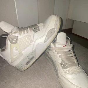 Nike Air Jordan 4 Retro White Cement - Säljer ett par Nike Air Jordan 4 Retro White Cement. De är vita med grå och svarta detaljer, meshpaneler och klassisk Jumpman-logga på hälen. Skorna har snörning, platt sula och är tillverkade i skinn och syntet. Perfekta för dig som gillar ikoniska sneakers med streetstyle-vibe.