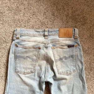 Ljusblå jeans från Nudie Jeans - Säljer ett par ljusblå jeans från Nudie Jeans med klassisk femficksdesign och snygga kontrastsömmar. Jeansen har raka ben och är tillverkade i ekologisk bomull. Bakfickorna har Nudie Jeans signaturbroderi och läderpatch i midjan. Hör av er vid frågor och funderingar!