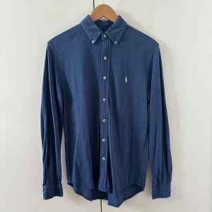 Blå skjorta från Polo Ralph Lauren - Snygg blå skjorta från Polo Ralph Lauren i lätt mesh-bomull. Klassisk button-down krage, vita knappar och broderad logga på bröstet. Perfekt för dig som gillar stilrena och tidlösa plagg med en sportig touch. Skriv för mer frågor och bilder