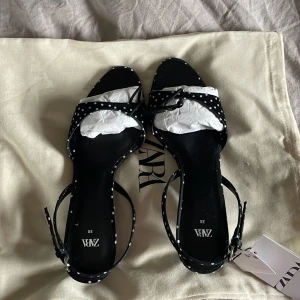 Prickiga svarta sandaler från Zara - Svarta sandaler från Zara med vita prickar, smal rem runt vristen och låg klack. Skorna har öppen tå och är tillverkade i ett glansigt syntetmaterial. Perfekt för dig som gillar retrovibbar och vill sticka ut med ett lekfullt mönster.