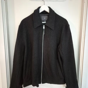 Mörkgrå ulljacka från H&M, M - Säljer en stilren mörkgrå jacka från H&M i ullmix. Jackan har klassisk krage, rak passform och dragkedja framtill. Perfekt för höst och vinter, med två fickor på sidorna och enkel design som funkar till det mesta.