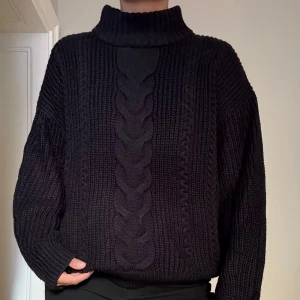 Svart stickad tröja med flätmönster - Mysig svart stickad tröja med grovt flätmönster framtill. Tröjan har en hög krage och är långärmad med en loose passform