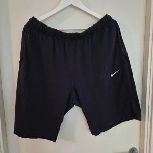 Svarta Nike shorts i bomull - Svarta shorts från Nike med klassisk vit swoosh-logga på ena benet. De har elastisk midja och är tillverkade i mjuk bomull, perfekta för chill dagar eller träning. Fickor på sidorna och en bakficka för extra funktionalitet.
