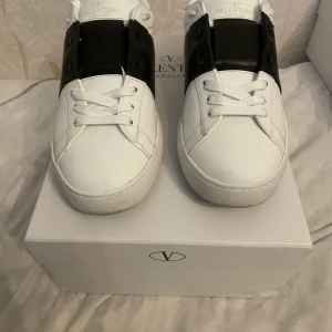 Valentino Garavani Open sneakers vit/svart - Snygga Valentino Garavani Open sneakers i vitt skinn med svart band över vristen och ikoniska nitar baktill. Klassisk rund tå, vita snören och platt sula. Kommer med originalkartong, dustbag och äkthetskort. Perfekta för dig som gillar lyxig streetstyle.