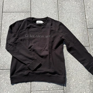 Svart sweatshirt från Calvin Klein Jeans - Svart sweatshirt från Calvin Klein Jeans med rund halsringning och diskret logga framtill. Tröjan har långa ärmar, ribbade muddar och är tillverkad i mjuk bomullsmix. Perfekt för en clean och stilren look.