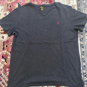 Mörkgrå v-ringad t-shirt Polo Ralph Lauren - Snygg mörkgrå t-shirt från Polo Ralph Lauren med v-ringad hals och korta ärmar. Klassisk rosa Polo-logga broderad på bröstet. Mjuk och skön bomull som passar perfekt till jeans eller shorts. Enkel och stilren design som funkar året runt.