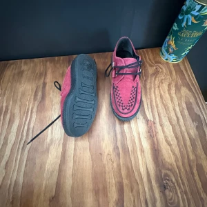 Röda creepers från T.U.K i mocka - Unika röda creepers från T.U.K med svart snörning och coolt flätmönster framtill. Ovandel i mocka och robust svart sula med döskalle- och benmotiv undertill. Perfekta för dig som vill sticka ut med din stil.