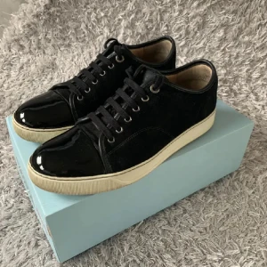 Svarta sneakers från Lanvin med lackdetalj - Snygga svarta sneakers från Lanvin med ovandel i mocka och glansig lackad tå. Skorna har vita sulor och klassisk snörning. Perfekt för dig som gillar stilrena men ändå unika detaljer på dina sneakers.