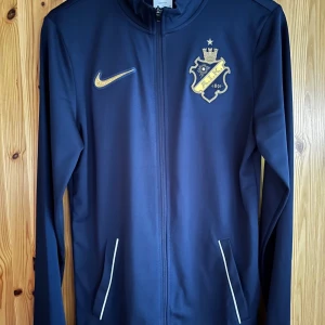 AIK NIKE Europa 2022 Tracksuit - Oanvänt - Storlek S - Ett matchande träningsset från Nike och AIK. Setet består av en träningsjacka med dragkedja och ett par träningsbyxor. Båda plaggen har AIK-märket och Nike-loggan.