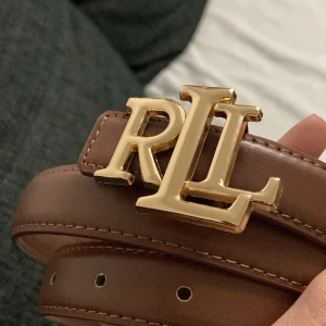 Brunt bälte, RLL design  - 🤎 Ett stilrent brunt bälte med guldigt RL-spänne framtill. Helt nytt och oanvänd. Bältet har en klassisk design som gör det enkelt att kombinera med bäde vardagskläder och mer uppklädda outfits. det finns flera häl för justerbar passform. Ett mängsidigt bälte som passar bäde till byxor, kjol eller för att markera midjan över en klänning.  Kontakta gärna vid frågor 🙏