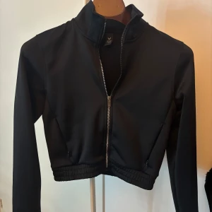 Svart croppad jacka från SOC XS - Svart croppad jacka från SOC med dragkedja framtill och hög krage. Jackan har långa ärmar, elastisk nederkant och två fickor med dragkedja. 