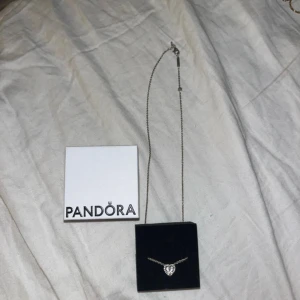 Pandora halsband med hjärthänge - Säljer ett snyggt halsband från Pandora i silver med ett glittrigt hjärthänge. Kedjan är tunn och har en diskret Pandora-tag vid låset. Perfekt för dig som gillar stilrena smycken med lite extra bling. Kommer med originalask. 