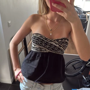 Svart bandeau tubtopp med broderi - Säljer en svart bandeau tubtopp med vitbroderat mönster upptill. Toppen är axelbandslös och har en luftig passform nedtill. Perfekt för varma dagar och enkel att styla med jeans eller kjol.