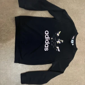 Svart Adidas sweatshirt med camomönster - Svart sweatshirt från Adidas med stort tryck på bröstet i form av Adidas-loggan med camouflagemönster i vitt, gult och lila. Tröjan har rund halsringning och långa ärmar. Perfekt för dig som gillar streetwear och sportig stil.