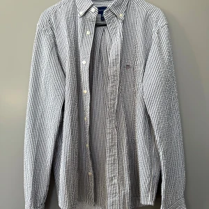 Randig skjorta från Gant i bomull - Säljer en klassisk randig skjorta från Gant i vitt och blått. Skjortan har lång ärm, knappar hela vägen och liten broderad logga på bröstfickan. Tillverkad i mjuk bomull och har en relaxed passform med button down-krage.