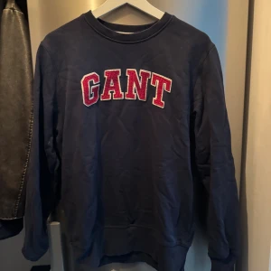 Mörkblå sweatshirt från GANT - Snygg mörkblå sweatshirt från GANT i storlek M (herrstorlek). Tröjan har ett stort broderat GANT-tryck i rött och vitt på bröstet. Klassisk rund hals och långa ärmar.