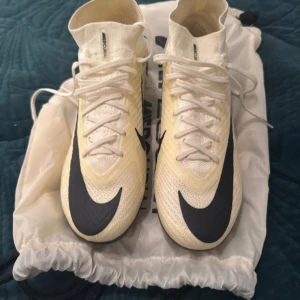 Nike Mercurial beiga fotbollsskor - Säljer ett par Nike Mercurial fotbollsskor i vitt med svarta detaljer och stor Swoosh-logga. Skorna har snörning, högre strumpliknande skaft och är tillverkade i lätt syntetmaterial för bästa känsla på planen. Perfekta för dig som vill sticka ut på matchen.