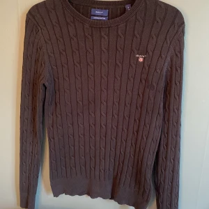 Mörkbrun kabelstickad tröja från GANT - Snygg mörkbrun kabelstickad tröja från GANT i stretchig bomull. Klassisk rund hals och broderad logga på bröstet. Perfekt för dig som gillar stilrena och tidlösa plagg med lite preppy känsla. Långärmad och med ribbade muddar för extra komfort.