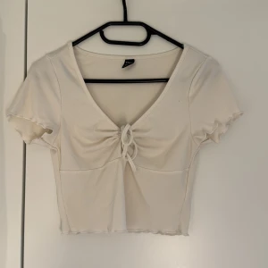 Vit ribbad croppad topp från SHEIN - Superfin vit croppad topp från SHEIN med ribbad struktur och söt knytning framtill. Toppen har korta ärmar med vågig kant och v-ringning. Perfekt för dig som gillar en enkel men ändå trendig stil. Den är lite urtvättad men annars inga fel. 