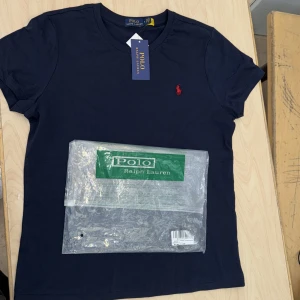 Mörkblå t-shirt från Polo Ralph Lauren - Snygg mörkblå t-shirt från Polo Ralph Lauren med klassisk röd broderad logga på bröstet. T-shirten har rund hals och korta ärmar, tillverkad i mjuk bomull som känns skön mot huden. Perfekt för dig som gillar stilrena och tidlösa plagg. storlek S i sverige men tröjan står i M