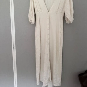 Lång vit klänning från NA-KD - Superfin långklänning från NA-KD i en cremevit färg och i hårdare linne material. Klänningen har djup v-ringning, knappar framtill och puffiga trekvartsärmar. Klänningen säljs då den inte används. Sömnen har gått av vid nacken men är något som inte syns och är enkelt att laga om man vill det. 