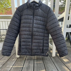 Svart pufferjacka från Everest - Svart pufferjacka från Everest med horisontella quiltade sömmar och hög krage. Jackan har dragkedja framtill och två sidofickor med dragkedja. Perfekt för kyliga dagar och har en clean, sportig look.